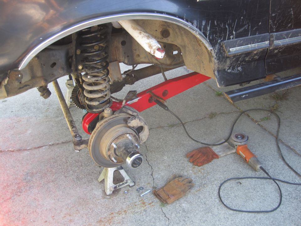 Close-up of vehicle's front suspension on jack stands, with grinder and gloves nearby