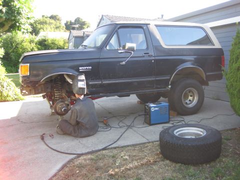1989 Ford Bronco - A person is welding on the front wheel assembly of a dark-colored Ford Bronco.