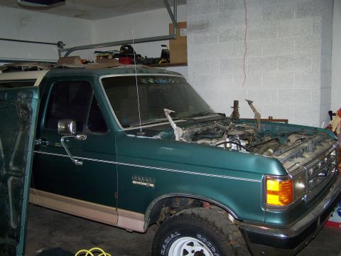 1989 Ford Bronco - A dark green 1989 Ford Bronco parked in a garage with its hood removed, exposing the engine bay; visible are the front left fender, side mi…