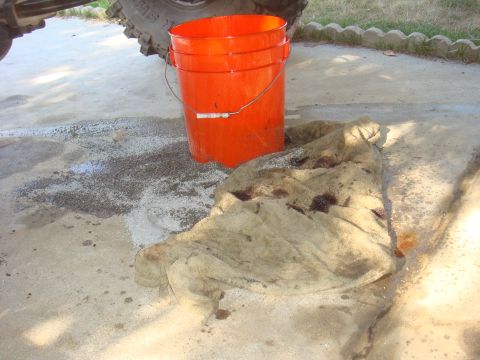 Orange bucket and stained rag next to a vehicle tire on a concrete surface with a spill
