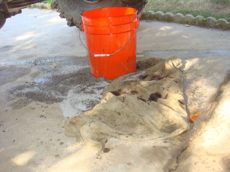 Orange bucket and stained rag next to a vehicle tire on a concrete surface with a spill