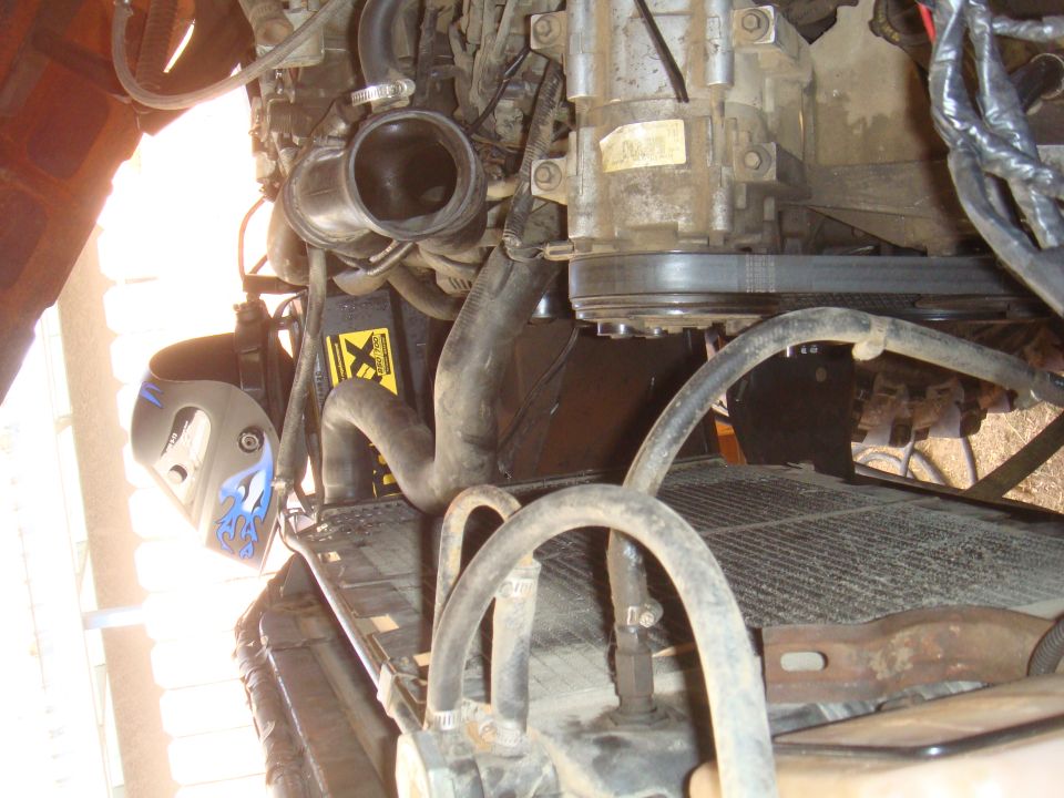 Engine bay with radiator, hoses, AC compressor, and belt visible. Yellow hazard sticker on battery