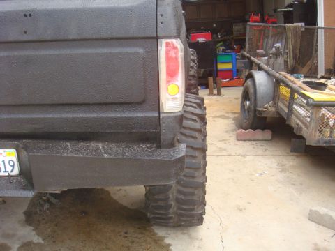 Rear of a black Ford Bronco with large off-road tires, parked next to a trailer in a garage