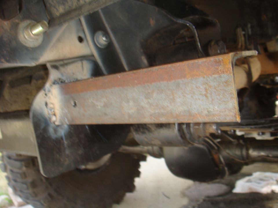 Close-up of rusted metal bracket welded to underside of vehicle frame, with tire visible