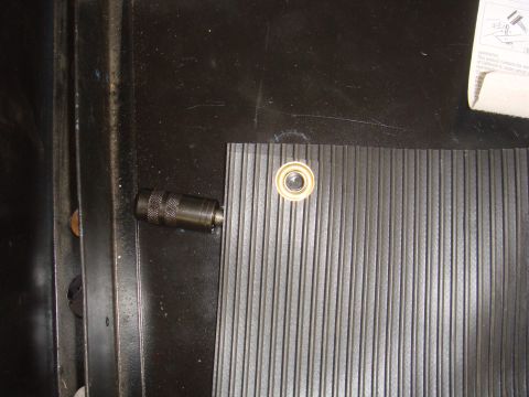 Close-up of a ribbed mat with a metal fitting and grommet in a dark vehicle interior. A document is partially