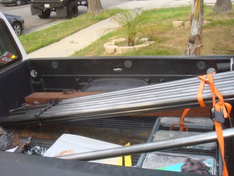 Truck bed interior with metal pipes secured by orange straps, a dark gray car and a truck visible in the backg