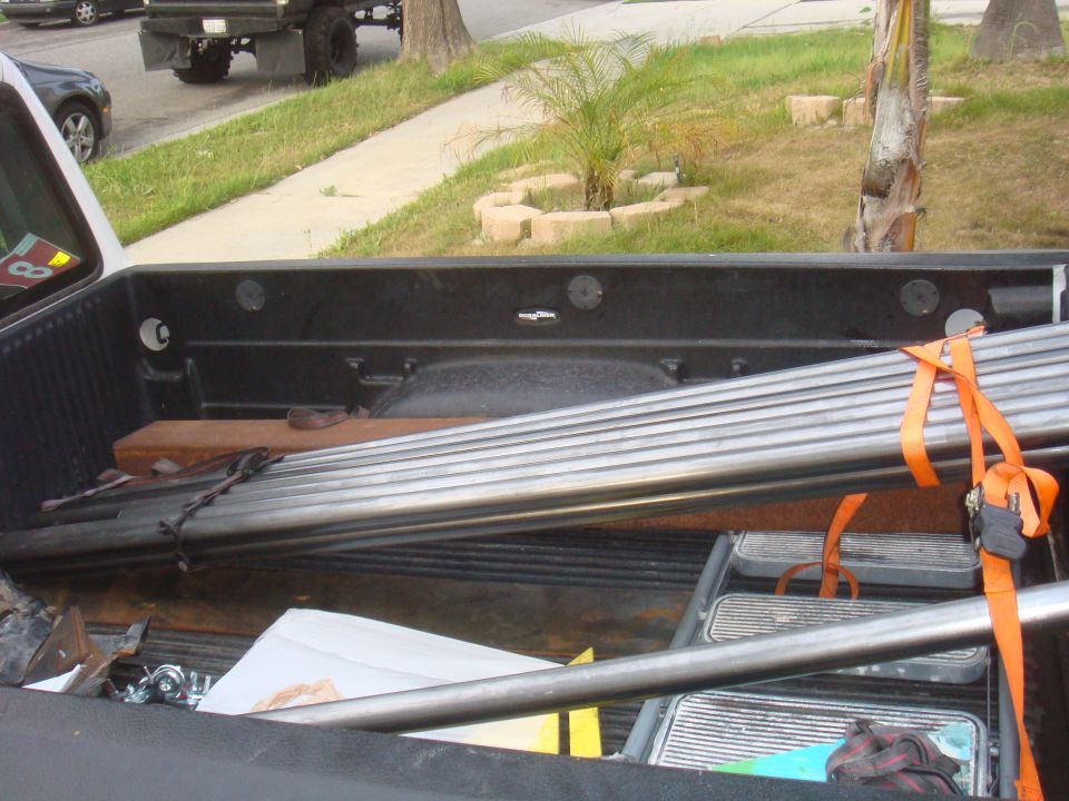 Truck bed interior with metal pipes secured by orange straps, a dark gray car and a truck visible in the background