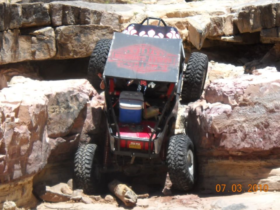 Off-road buggy with large tires navigating a rocky incline. A cooler is visible in the back