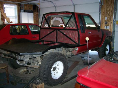 1988 Suzuki Samurai - A red pickup truck with a custom flatbed and roll cage is parked in a garage next to other vehicles.