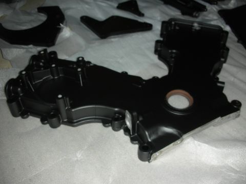 1992 Ford Crown Victoria - A black engine timing cover is shown from an elevated angle, with other black engine parts scattered in the background.