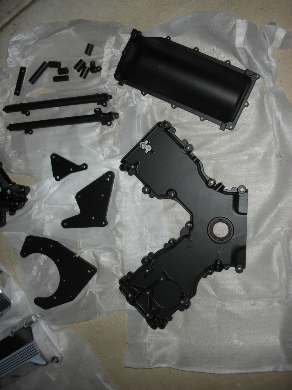 Black engine timing cover and other assorted engine parts laid out on white paper