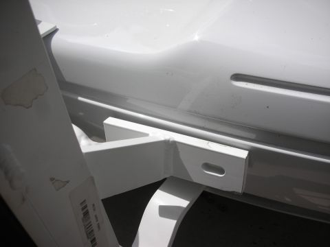 2004 Ford Crown Victoria - Close-up under the rear of a white sedan showing a white-painted welded tube trailer hitch receiver bracket with a slotted mounting tab, tu…