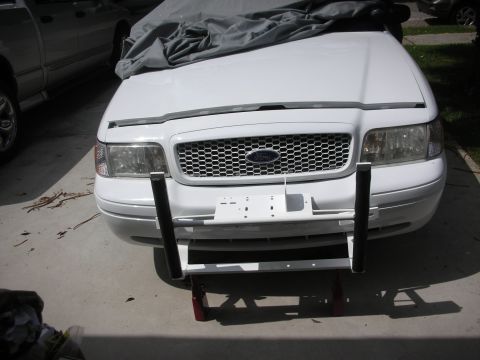 2004 Ford Crown Victoria - Front view of a white Ford Crown Victoria sedan on a sunny concrete driveway, wearing a clear hood deflector and a white push bumper whose…