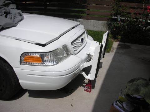 2004 Ford Crown Victoria - Front corner of a white Ford Crown Victoria in a driveway mid-install of a white push bumper, with the upright and mounting brackets proppe…
