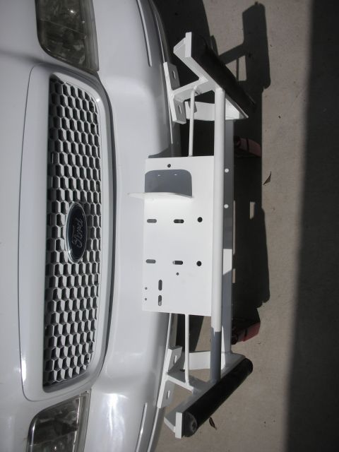2004 Ford Crown Victoria - Rotated close-up of a white-painted police-style push bumper propped in front of a white Ford sedan's honeycomb grille and blue Ford oval b…