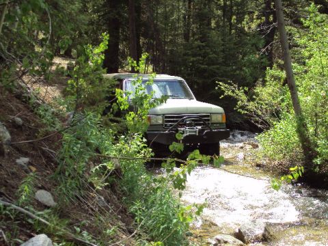 1989 Ford Bronco - A green Ford Bronco navigates a rocky stream bed surrounded by dense foliage.