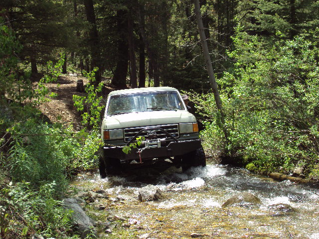 White 1989 Ford Bronco driving through a stream in a forest