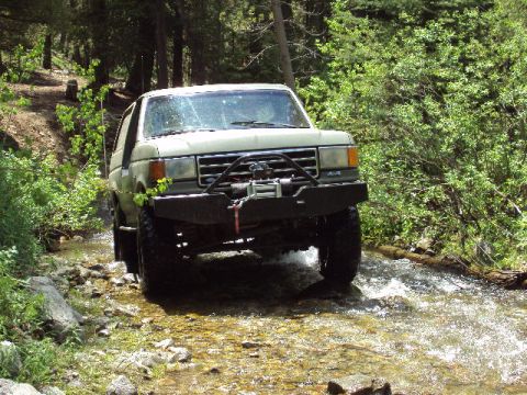 1989 Ford Bronco - 1989 Ford Bronco navigates a rocky stream bed surrounded by trees.