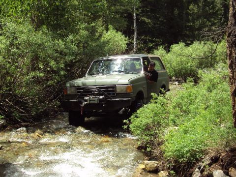1989 Ford Bronco - This Ford Bronco is navigating a stream in a forest, with lush green foliage surrounding it.