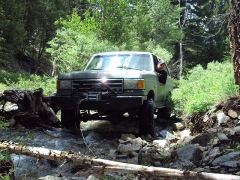 1989 Ford Bronco - A green Ford Bronco navigates a challenging rocky stream bed surrounded by dense forest.