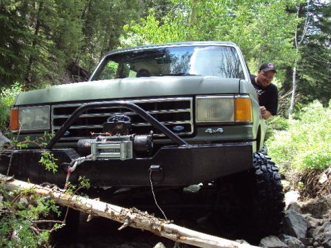 1989 Ford Bronco - This green Ford Bronco, likely from the late 1980s, navigates a rocky, wooded trail with a winch mounted on its bull bar.