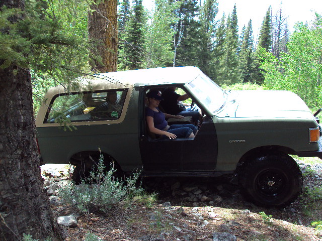 Green Ford Bronco with tan top, door open, off-road tires, in wooded area