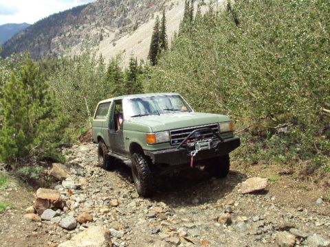 1989 Ford Bronco - This SUV is navigating a rough, rocky mountain path surrounded by trees and steep terrain.