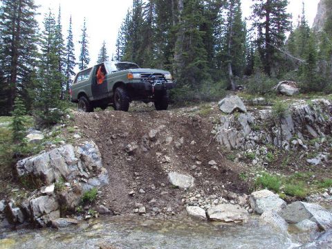1989 Ford Bronco - A green Ford Bronco navigates a steep, rocky incline in a forested mountain area.