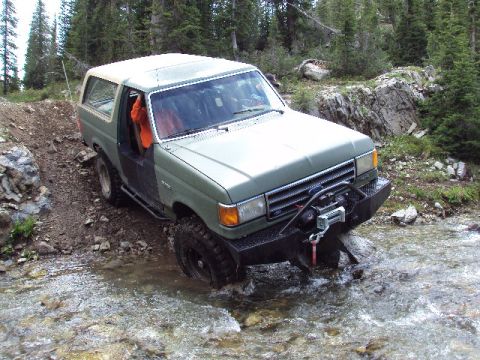 1989 Ford Bronco - This Ford Bronco navigates a challenging off-road stream crossing amidst a wooded mountain landscape.