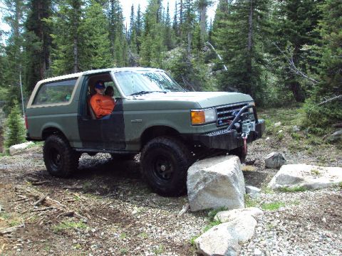 1989 Ford Bronco - This Ford Bronco is shown off-roading on a rocky trail surrounded by trees.