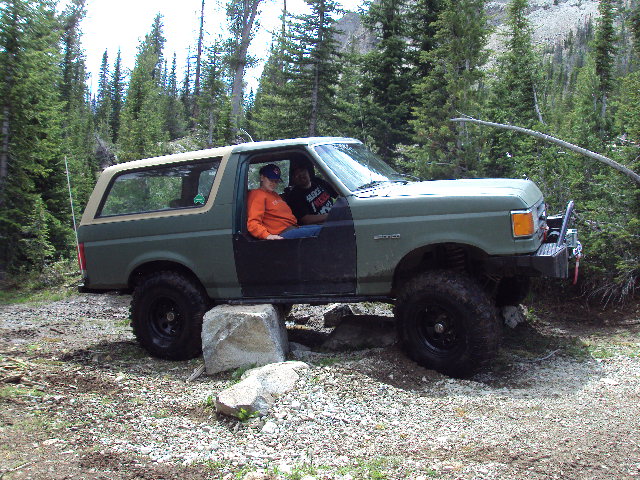 1989 Ford Bronco with two people inside, driving over rocks in a forest