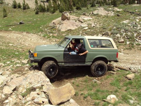 1989 Ford Bronco - A green Ford Bronco with a tan top is shown off-roading on a rocky mountain path.