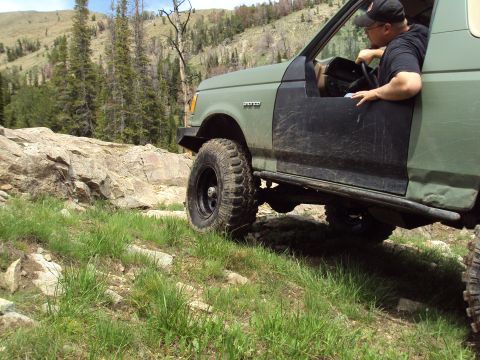 1989 Ford Bronco - This 1989 Ford Bronco is shown off-roading on a rocky, grassy slope with trees and hills in the background.