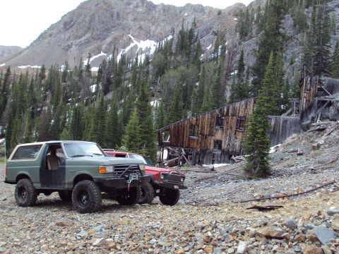 1989 Ford Bronco - Two off-road vehicles parked near a derelict wooden structure in a mountain setting.