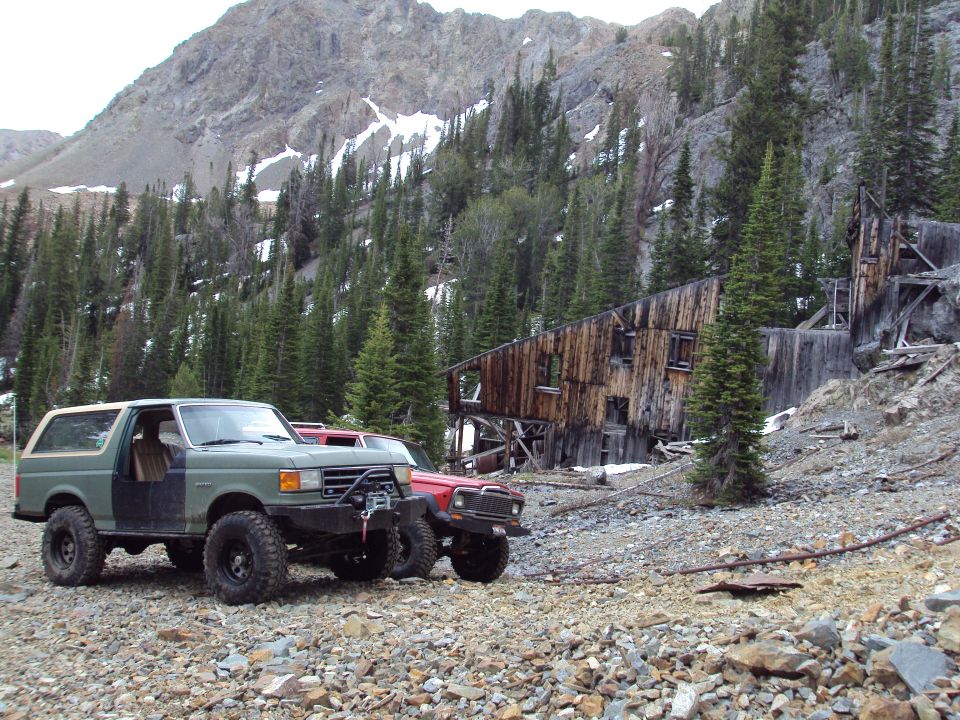 Green 1987 Ford Bronco with winch and red Jeep Cherokee parked on rocky ground near abandoned wooden building