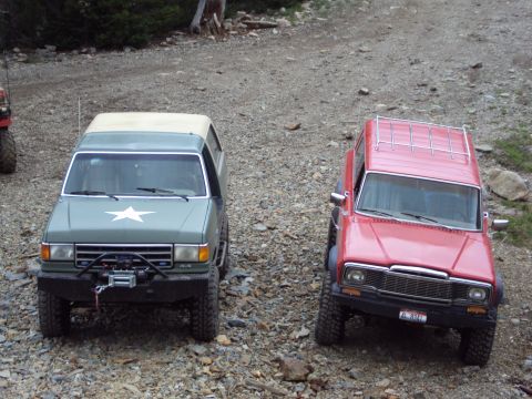 1989 Ford Bronco - Two modified SUVs, one green and one red, are parked on a rocky incline in a wooded area.