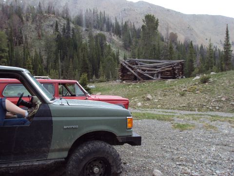 1989 Ford Bronco - A green 1989 Ford Bronco and a red SUV are parked near a rustic cabin in a mountainous, wooded area.