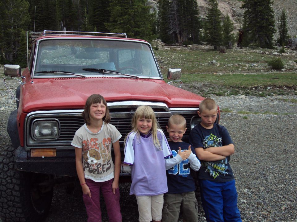 Four children stand in front of a red SUV with a roof rack. The SUV has a prominent grille and large tires