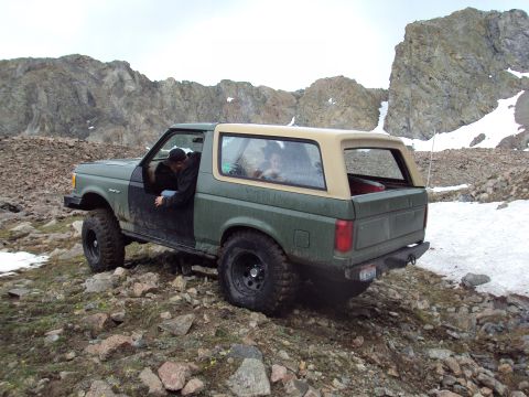 1989 Ford Bronco - A green Ford Bronco with a tan top navigates a rocky, mountainous landscape with patches of snow.