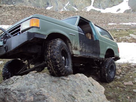 1989 Ford Bronco - Green Ford Bronco with aftermarket bumper and large tires climbing over rocks in a mountainous area.