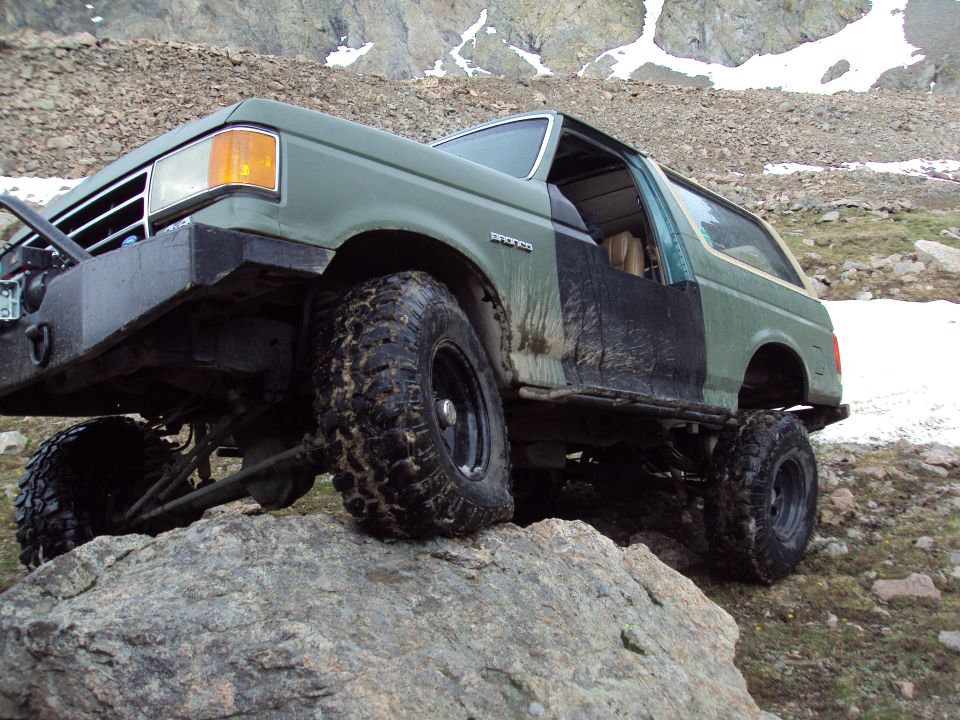 Green Ford Bronco with aftermarket bumper and large tires climbing over rocks