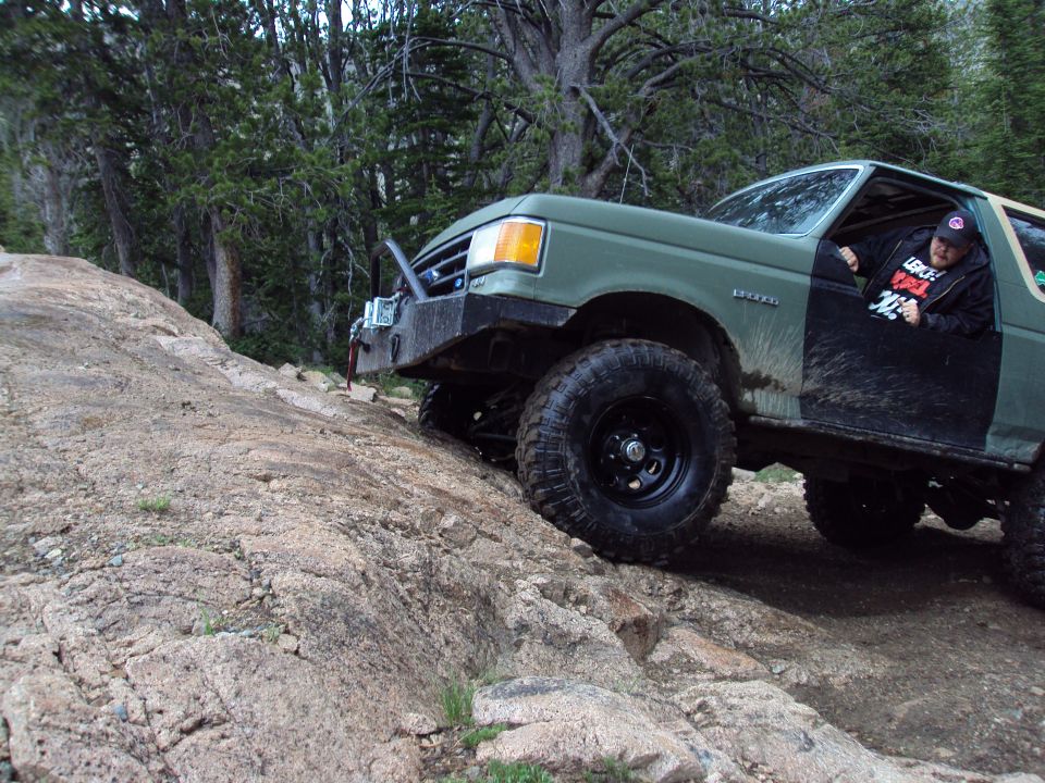 Green Ford Bronco climbing rocky terrain. Driver wearing a black hat and jacket