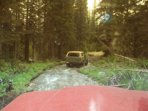 1989 Ford Bronco - A green SUV navigates a stream in a dense forest, with the front of another vehicle visible in the foreground.