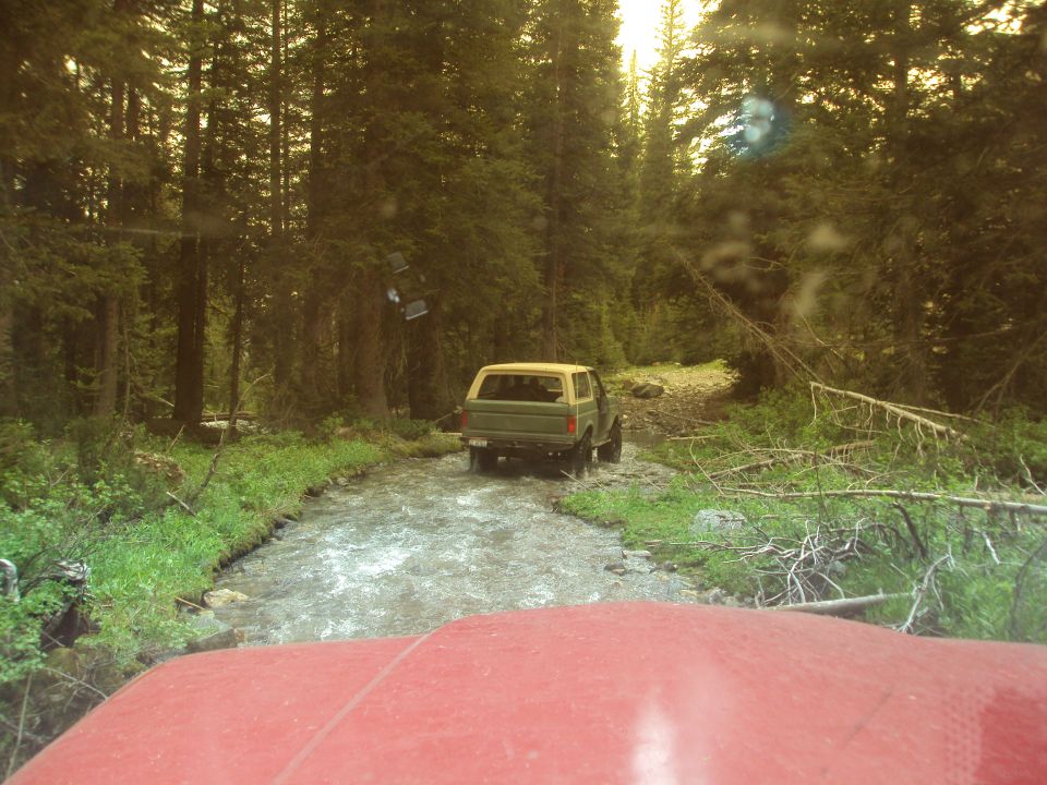 Green SUV driving through a stream in a forest, seen from the red hood of another vehicle