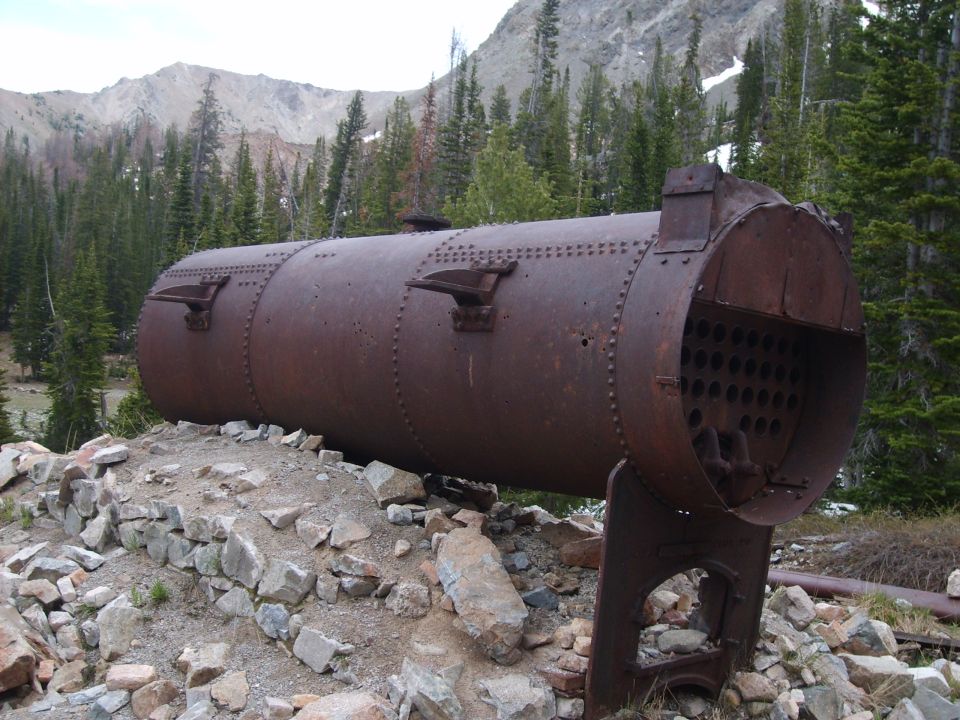 Rusty boiler on rocky ground with trees and mountains behind