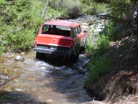 1989 Ford Bronco - A red SUV navigates a rocky stream bed surrounded by lush green foliage.