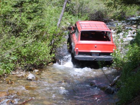 1989 Ford Bronco - Off-road vehicle navigates a rocky stream bed surrounded by dense green foliage.