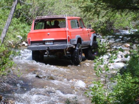 1989 Ford Bronco - This red Jeep Cherokee is navigating a rocky stream bed surrounded by lush green foliage.
