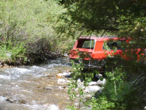1989 Ford Bronco - A red SUV navigates a shallow, rocky stream surrounded by dense foliage.