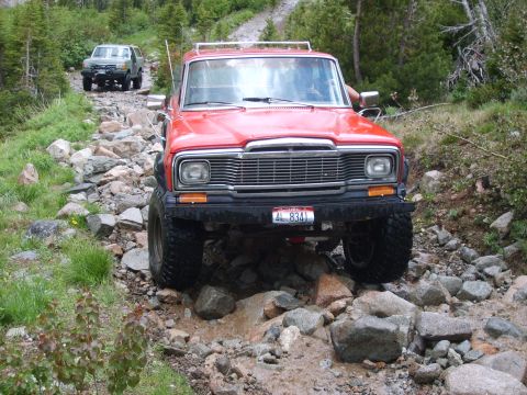 1989 Ford Bronco - A red Jeep Wagoneer drives over a rocky path, with another SUV following behind on the trail.